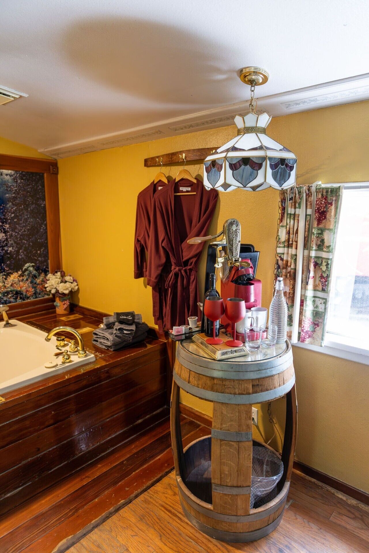 A bathroom with a wooden bathtub, a round wooden stool with a glass top, a red robe hanging on a rack, and a decorative ceiling light.