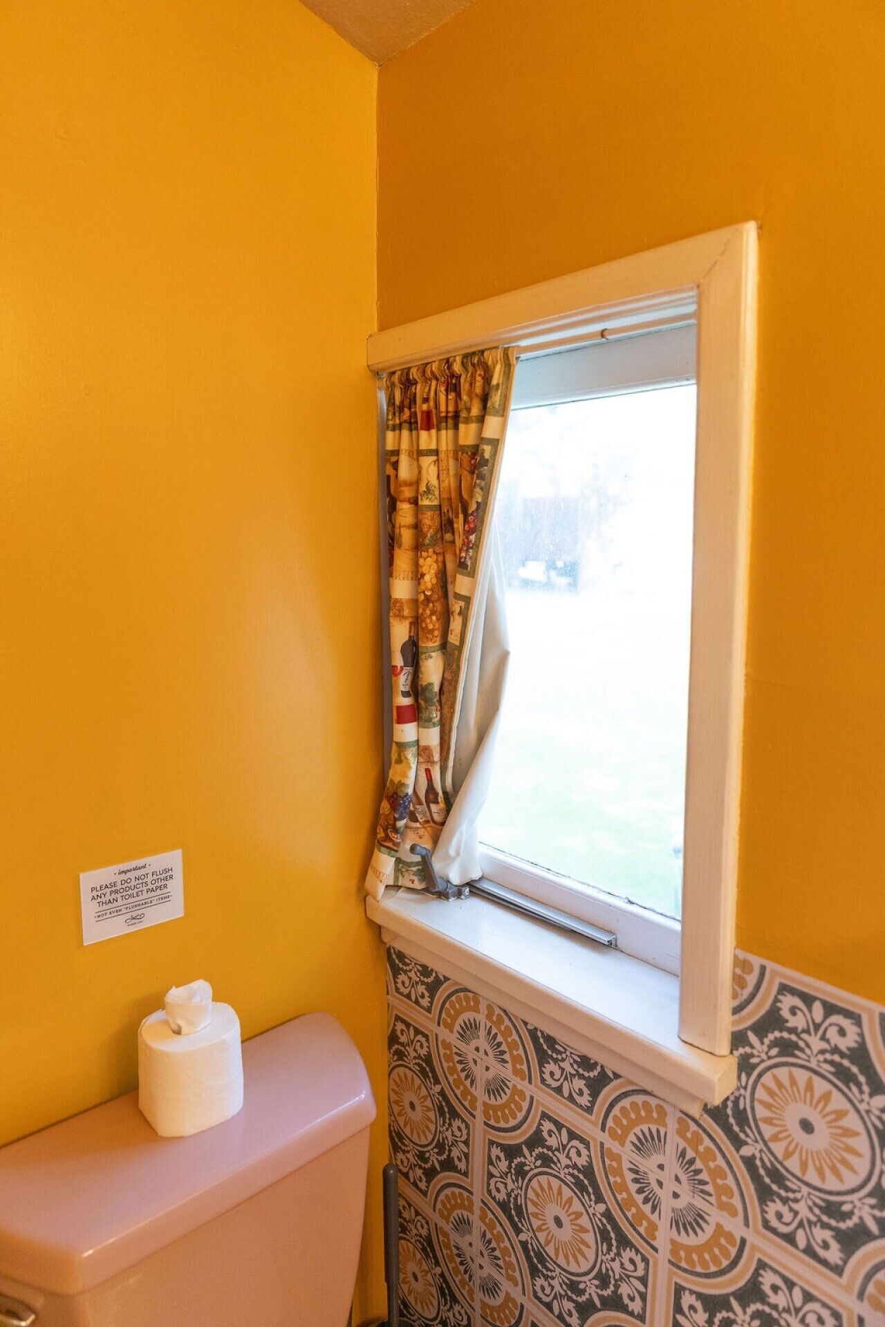 Een badkamer met toilet, toiletpapier en een raam met gordijnen met patroon.