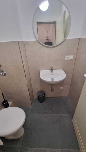 A compact bathroom with a white sink, a toilet, a round mirror, and a trash bin.