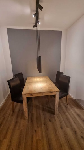 A dining area with a wooden table and two chairs, a modern pendant light, and a plain wall.