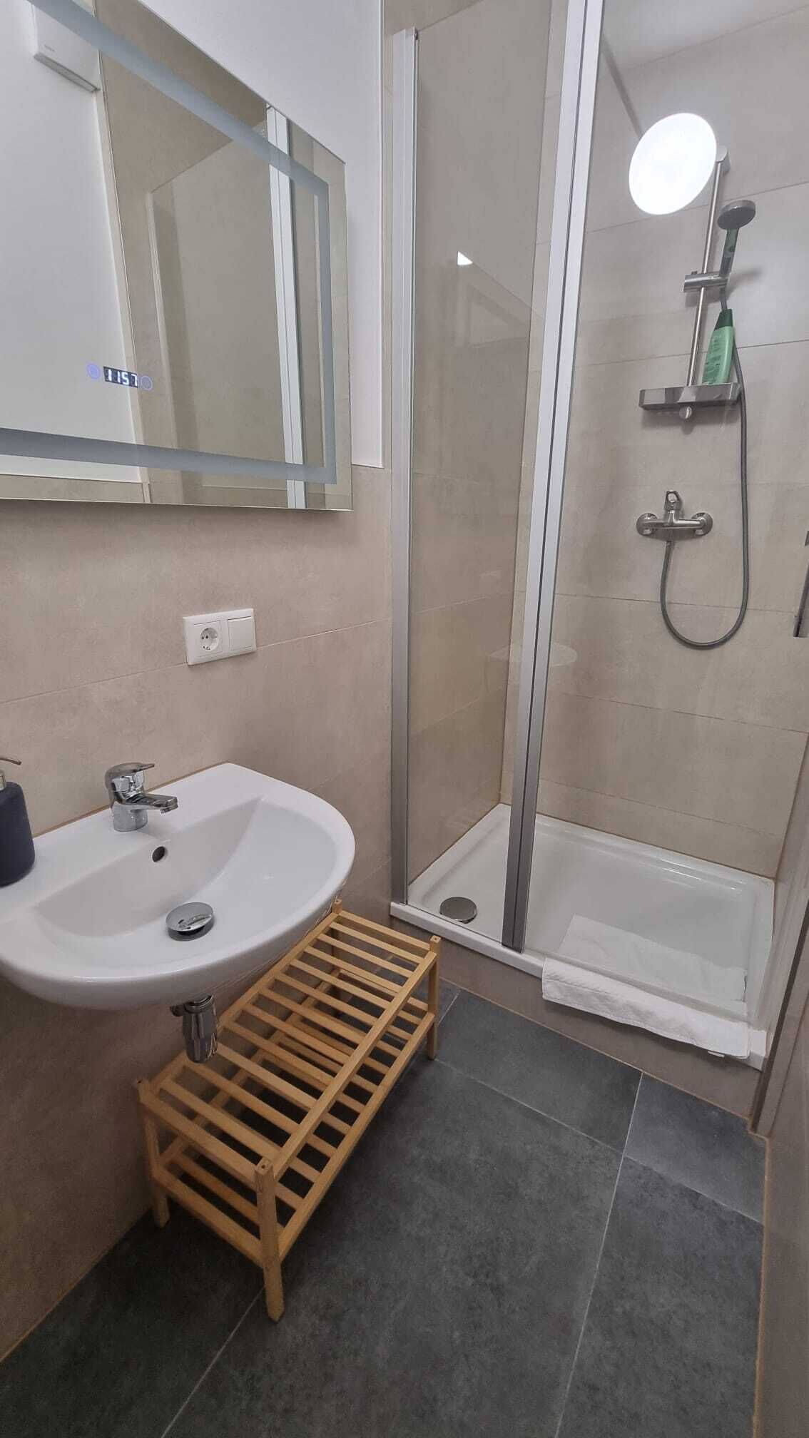 A bathroom with a white sink, a mirror, a shower with a glass door, and a wooden shelf.