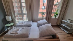 En dobbeltseng med hvide sengetøj, et natbord med lampe, og et vindue med udsigt til en murstensbygning.