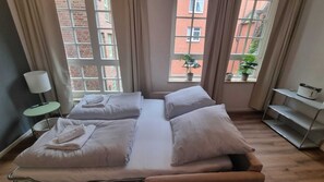 Ein Doppelbett mit weißer Bettwäsche, ein Nachttisch mit Lampe und ein Fenster mit Blick auf ein Backsteingebäude.