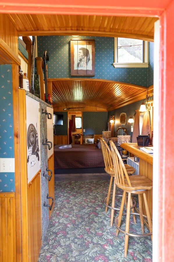 A compact room with a bed, a desk with chairs, a small window, and a wall-mounted picture.