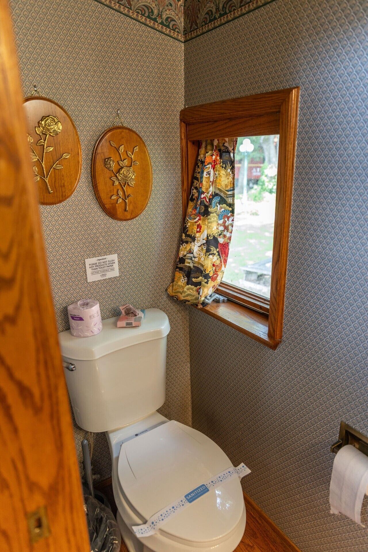 Een badkamer met een toilet, een houten plaquette met een bloemenmotief en een raam met een gordijn met dessin.