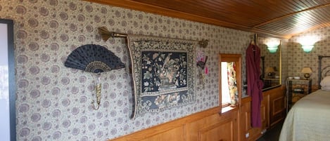 A room with patterned wallpaper, a wooden ceiling, a bed, a bedside table, a mirror, and a hanging bathrobe.
