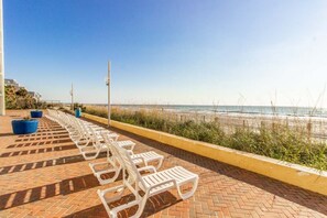 A paved walkway with white lounge chairs and blue planters, leading to a beach with a clear view of the ocean. - The Coastal SM | Fifty Oh Four Oh Three (Myrtle Beach)