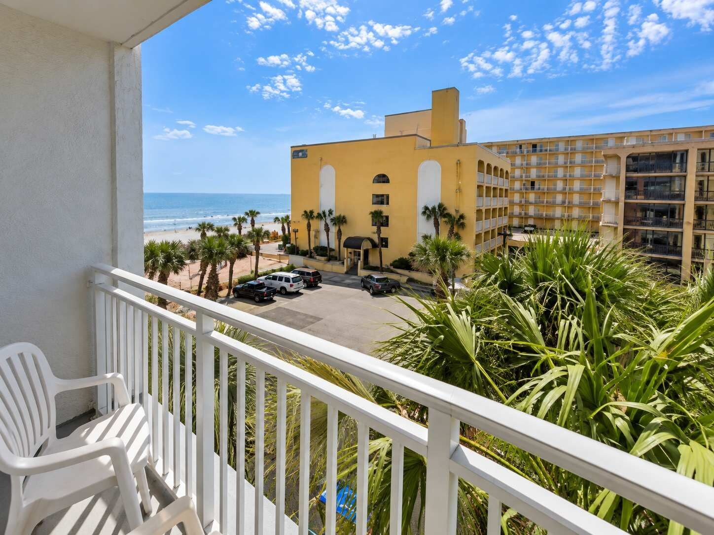 Un balcone con ringhiere bianche che si affaccia su un'area parcheggio, un edificio con diversi balconi e vista sulla spiaggia.