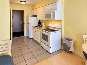 A compact kitchen with white appliances, including a refrigerator, microwave, and oven, and a trash bin. - The Coastal SM | Fifty Oh Four Oh Three (Myrtle Beach)