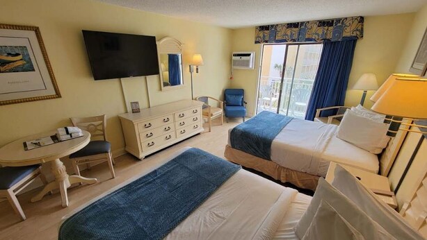 Habitación de hotel con dos camas, un escritorio, una silla, un aparador, televisor y balcón con vistas.