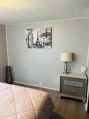 A bedroom with a bed, a nightstand with a lamp, a wall-mounted picture of the Eiffel Tower, and a power outlet. - 2-bedroom Zen minimalist mobile home by beaches and more! (Jacksonville)