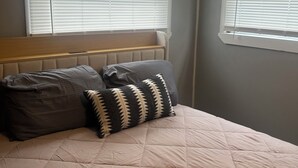 Un dormitorio con cama, dos almohadas, cabecera y dos ventanas con persianas.
