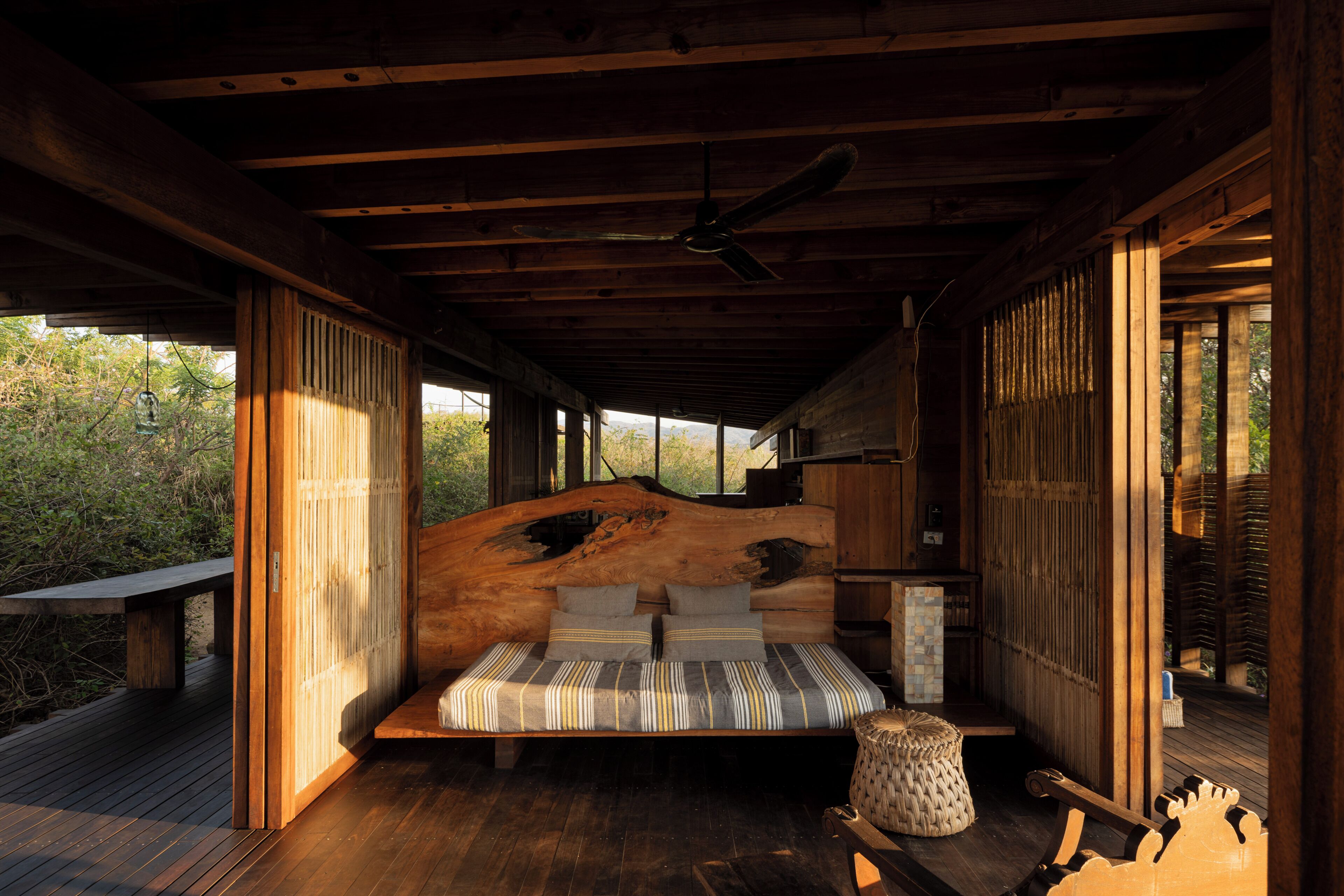 Interior de una cabaña de madera con una cama, un ventilador de techo y vistas al exterior.