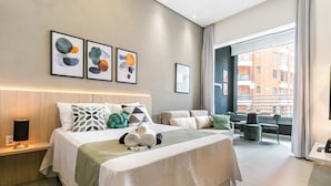 A modern hotel room with a large bed, a bedside table with a lamp, a view of the city through the window, and artwork on the wall. - Conforto prox metrô e Estacionamento (São Paulo)