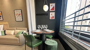 A modern living room with a sofa, a dining table set with plates and glasses, green chairs, and a view through a window with a railing. - Conforto prox metrô e Estacionamento (São Paulo)