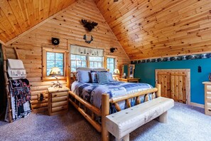 Ein Holzblockhaus-Interieur mit einem Bett, einer Bank, einem Schminktisch und einem Fenster mit Blick auf Bäume.