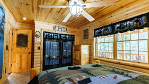 Een slaapkamer in een blokhut met een bed, een plafondventilator, een raam met gordijnen en een deur.