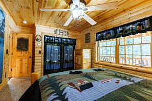 A log cabin bedroom with a bed, a ceiling fan, a window with curtains, and a door. - Bear-Themed Lodge / Sleeps 10 / Private Beach (Millersburg)