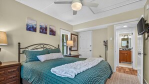 Ein Schlafzimmer mit einem Bett, Nachttischen, einem Deckenventilator, Kunstwerken an der Wand und einer Tür zu einem Badezimmer.