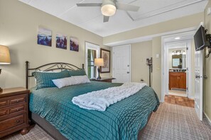 A bedroom with a bed, nightstands, a ceiling fan, artwork on the wall, and a door leading to a bathroom. - Pinnacle B09 (Killington)