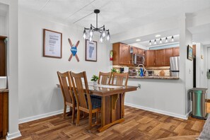 A dining area with a wooden table and chairs, a kitchen with wooden cabinets, and a hanging light fixture. - Pinnacle B09 (Killington)