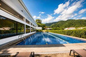 A modern pool with clear blue water, surrounded by a white building with large windows, and a view of green hills under a blue sky. - Modern Lakeview Penthouse: Pool/Gym/Restaurant (Río Oro)