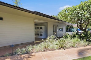 A single-story building with a covered porch, a garage with storage cabinets, and a landscaped garden with various plants. - Mahina Surf 123 & 124 (Lahaina)