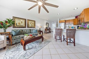 A spacious living room with a sofa, coffee table, and chairs, a kitchen area with wooden cabinets, and a ceiling fan. - Mahina Surf 123 & 124 (Lahaina)