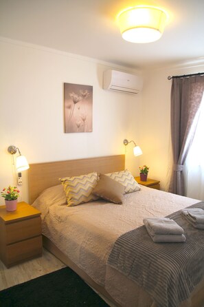 Ein ordentlich bezogenes Bett mit Kissen, einer Nachttischlampe, einer Wand-Klimaanlage und einem gerahmten Bild an der Wand.