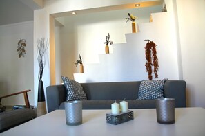 Ein modernes Wohnzimmer mit einem grauen Sofa, zwei gemusterten Kissen, einem Couchtisch mit Kerzen und Zimmerpflanzen.