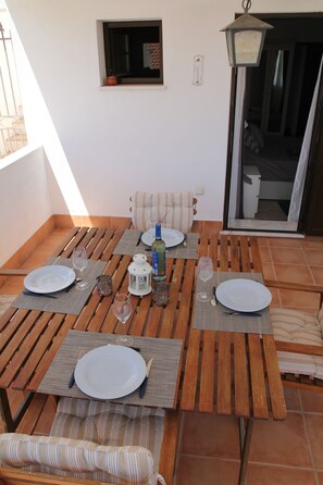 Ein Außenbereich mit Essplatz, Holztischen und gestreiften Kissen, einem kleinen Fenster und einer Tür, die auf einen Balkon führt.