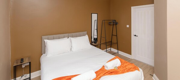 A neatly made bed with white bedding and an orange blanket, a black hanger rack, a mirror, and a white door.