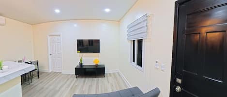 A modern living room with a flat-screen TV, a sofa, a dining area with a table and chairs, and a black door.