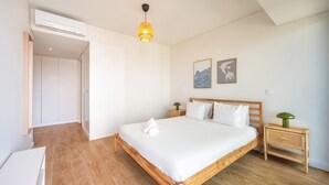 Ein Schlafzimmer mit einem hölzernen Bett, weißer Bettwäsche, einer Nachttischlampe, einer wandmontierten Klimaanlage und zwei gerahmten Kunstwerken.
