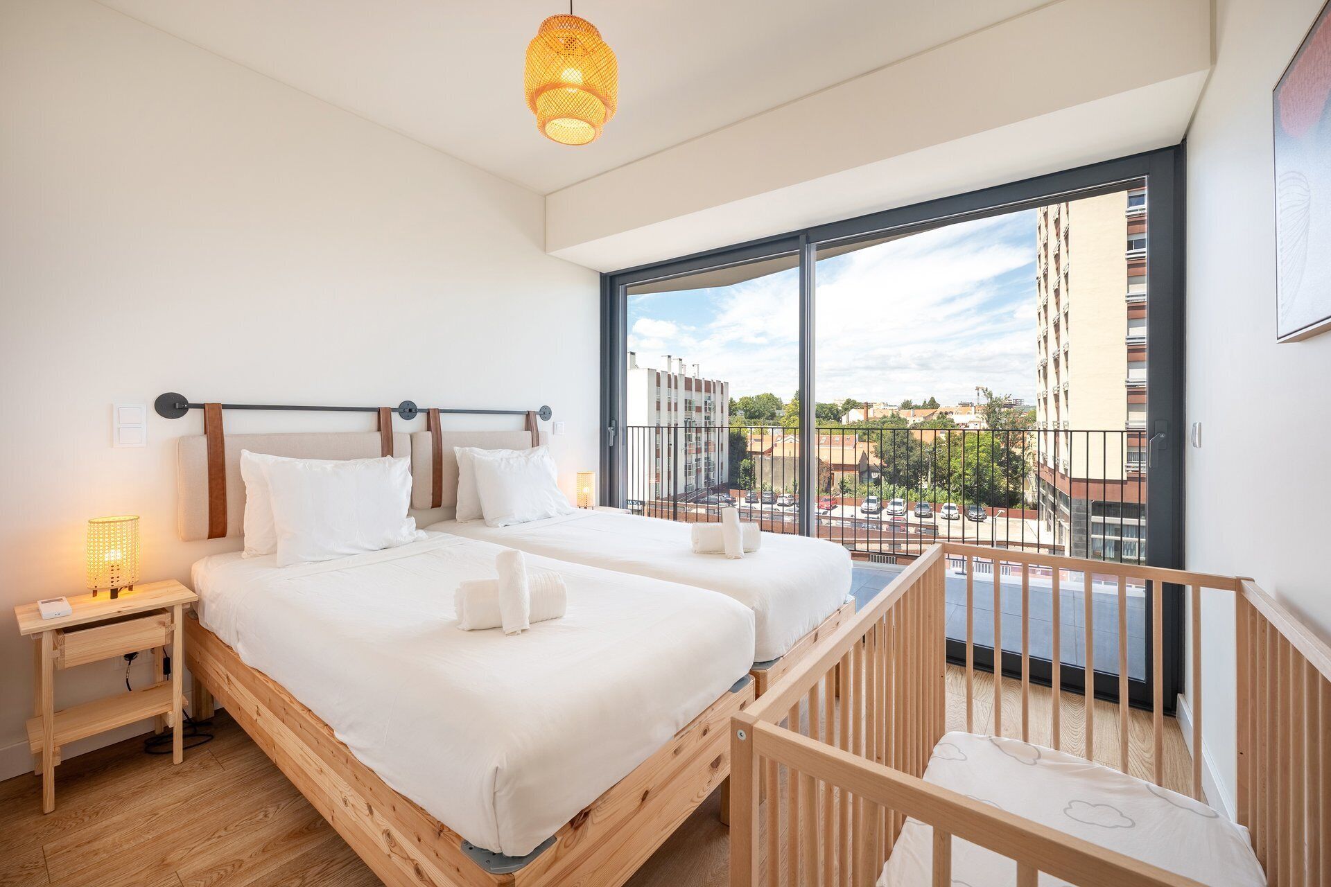 A modern bedroom with two beds, a wooden floor, a balcony with a railing, and a large window offering a city view.