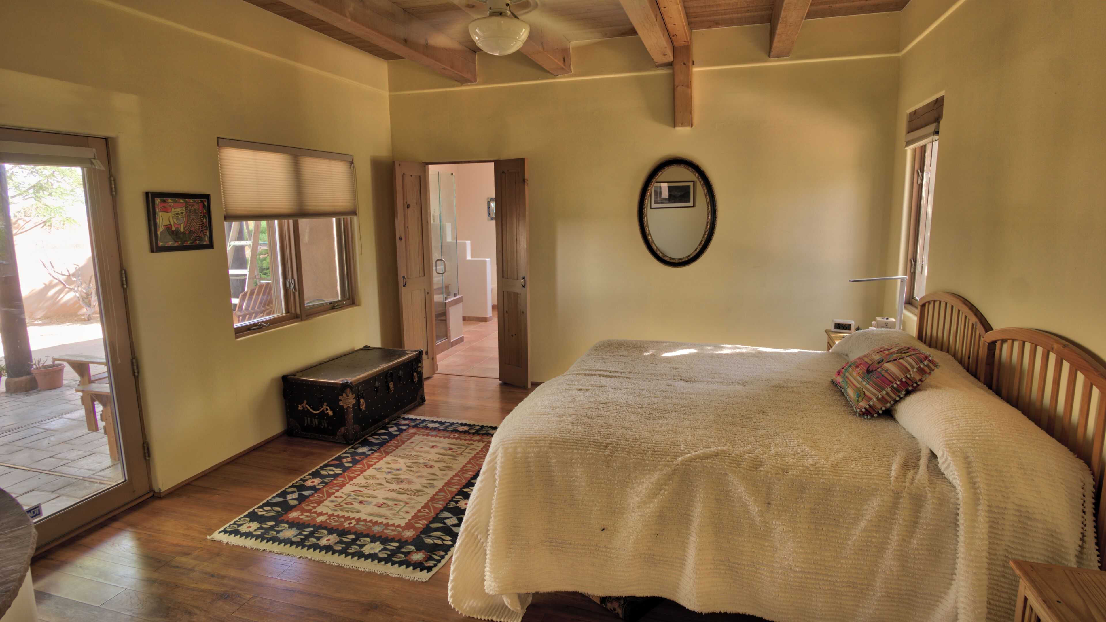 A bedroom with a large bed, a wooden headboard, a bedside table, a nightstand, a mirror, a window with blinds, a door leading to a staircase, and a suitcase on the floor.