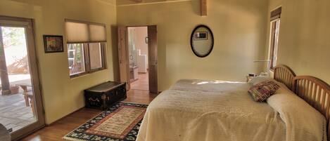 A bedroom with a large bed, a wooden headboard, a bedside table, a nightstand, a mirror, a window with blinds, a door leading to a staircase, and a suitcase on the floor.