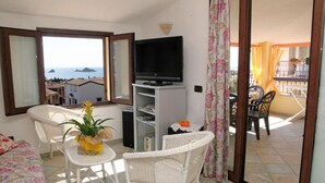 Een balkon met een televisie, rieten meubels, een kleine tafel met een plant en uitzicht op zee.