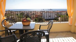 Een balkon met een tafel, stoelen, een mand vol fruit en uitzicht op zee en gebouwen.