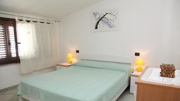 Een slaapkamer met een bed, twee nachtkastjes, een lamp op elk kastje, een schilderij aan de muur en een raam met gordijnen.