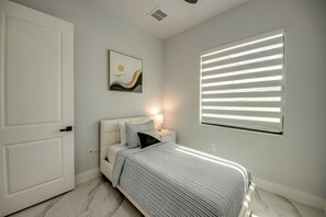 Un dormitorio con cama, mesita de noche, lámpara, ventana con persianas y puerta.