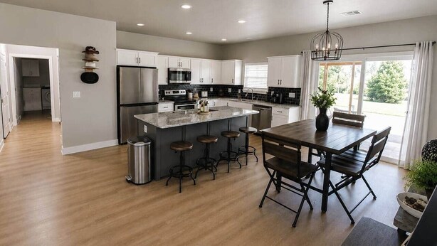 A modern kitchen with a central island, bar stools, and a dining table with chairs. - The Rock House | Walk to OSU (Stillwater)