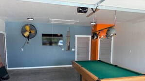 A pool table in a room with a ceiling fan, a mounted TV, and a door. - The Rock House | Walk to OSU (Stillwater)