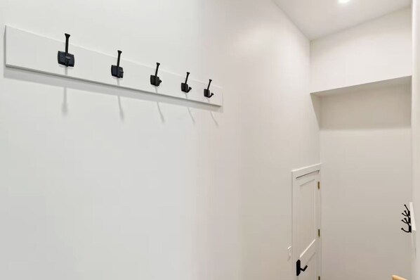 A hallway with a white wall, a door with a black handle, and a white shelf with black hooks.