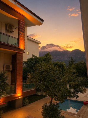 Un edificio moderno de dos pisos con balcón, piscina y vistas a las montañas al atardecer.