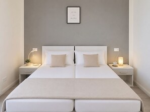 Un lit double avec une tête de lit blanche, deux tables de chevet avec des lampes et une citation encadrée au mur.