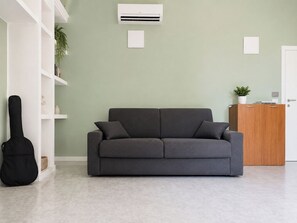 Ein Zimmer mit grauem Sofa, einer weißen Wandklimaanlage, einem Holzschrank und einem schwarzen Gitarrenkoffer.