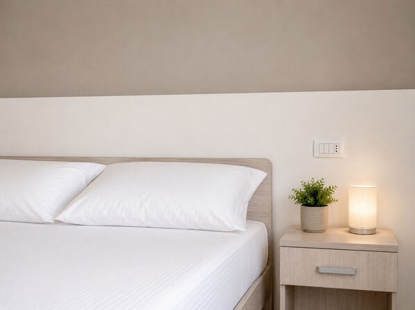 Ein ordentlich bezogenes Bett mit weißen Bettwäsche, ein Nachttisch mit Lampe und Topfpflanze sowie ein Lichtschalter an der Wand.
