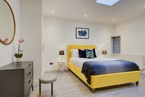 Une chambre à coucher comprenant un lit, des tables de chevet, un miroir, un petit tabouret et une fenêtre avec des stores.