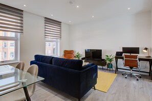 A modern living room with a dark blue sofa, a glass dining table, a TV, a desk with a computer, and a chair. - Hybrid Resi - Fulham - Three-Bedroom Apartment (London)